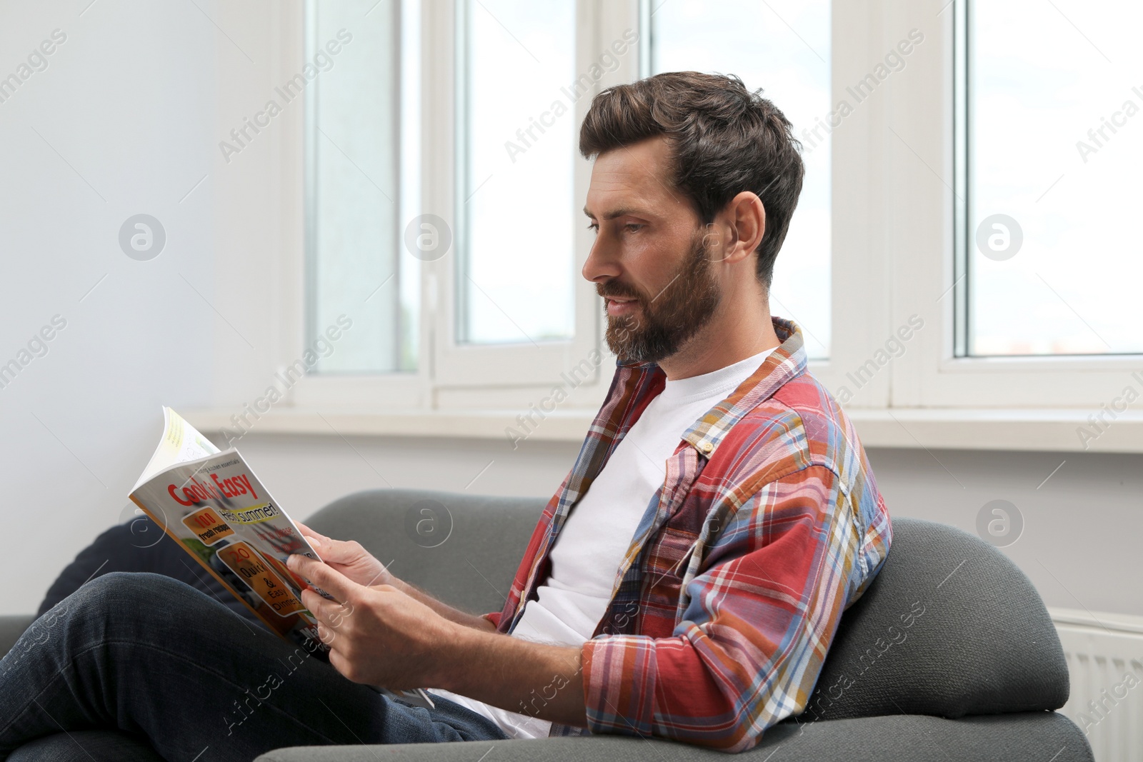 Photo of Smiling bearded man reading magazine on sofa indoors