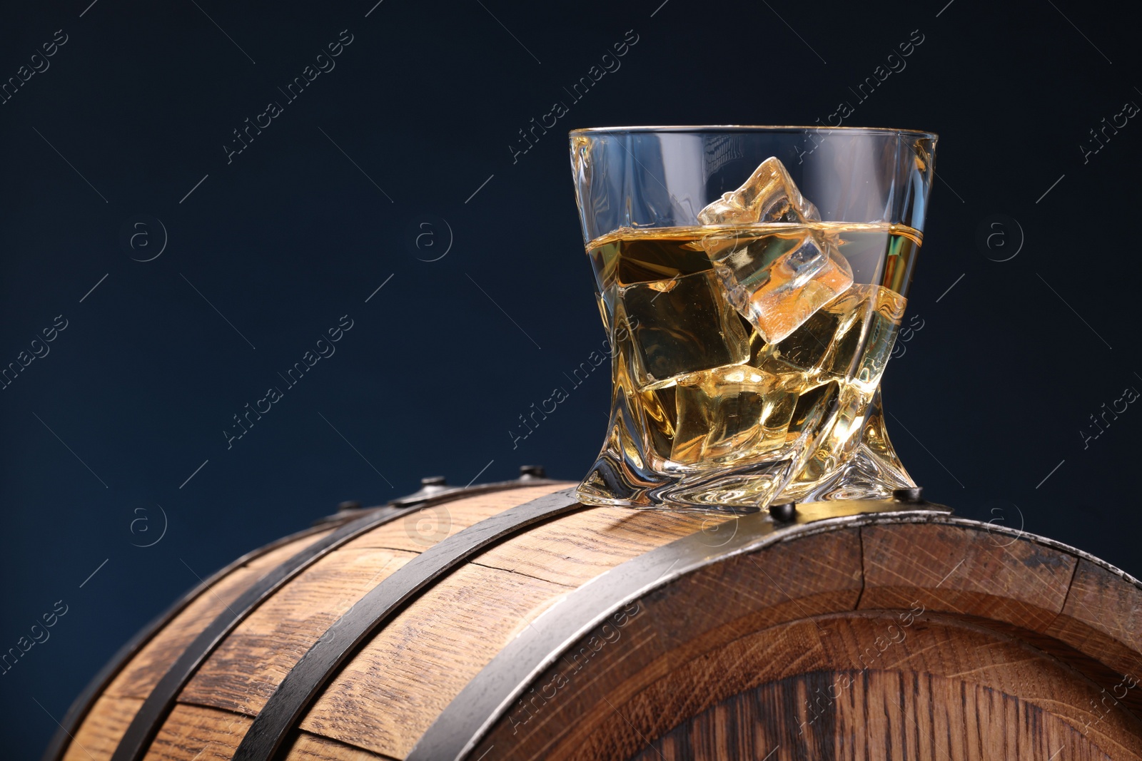 Photo of Whiskey with ice cube in glass on wooden barrel against blue background, space for text