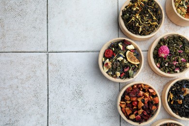 Different kinds of dry herbal tea in wooden bowls on white tiled surface, flat lay. Space for text