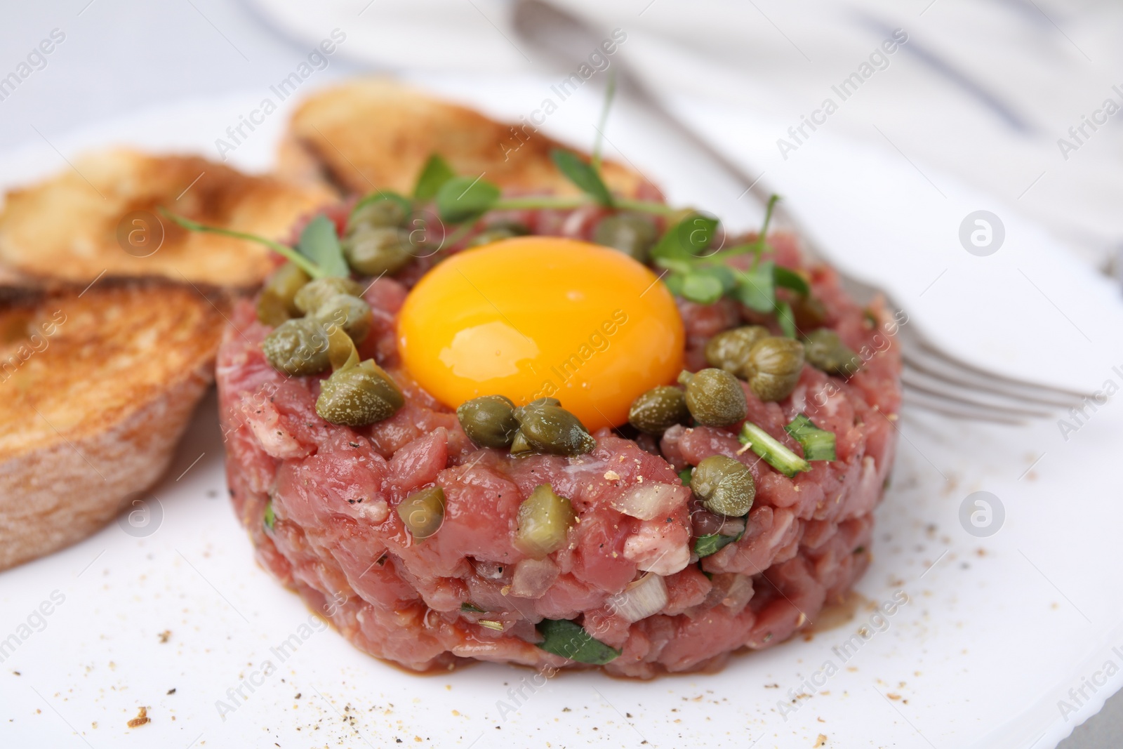 Photo of Tasty beef steak tartare served with yolk, capers, toasted bread and greens on plate, closeup