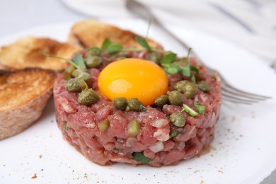 Photo of Tasty beef steak tartare served with yolk, capers, toasted bread and greens on plate, closeup
