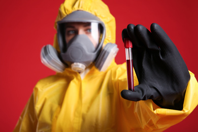 Photo of Woman in chemical protective suit holding test tube of blood sample against red background, focus on hand. Virus research