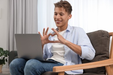 Photo of Happy young man having video chat via laptop and making heart with hands on armchair indoors. Long-distance relationship