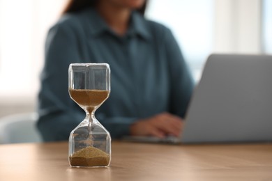 Photo of Hourglass with flowing sand on table. Woman using laptop indoors, selective focus