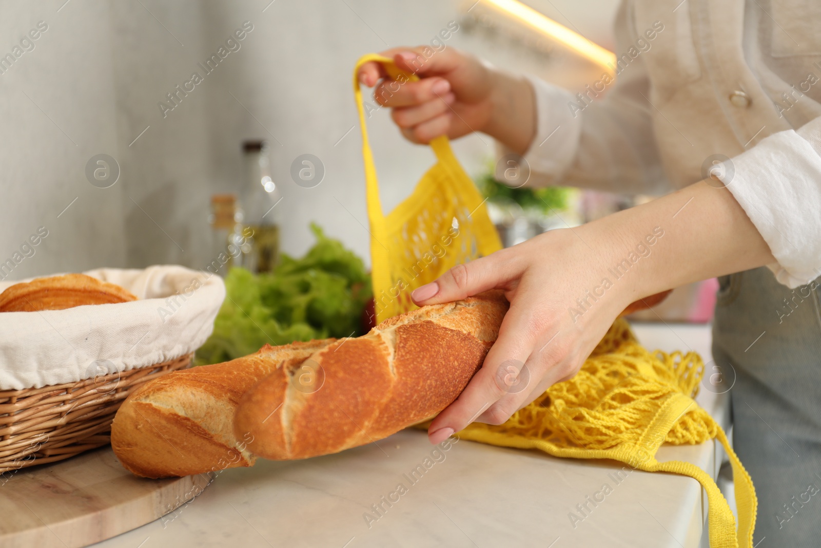 Photo of Woman taking baguette out from string bag at countertop, closeup