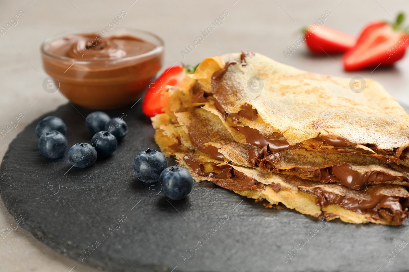 Photo of Delicious thin pancakes with chocolate spread and blueberries on grey table, closeup