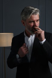 Handsome bearded man smoking cigar near dark grey wall indoors
