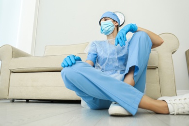 Photo of Exhausted doctor sitting on floor indoors, low angle view. Stress of health care workers during COVID-19 pandemic