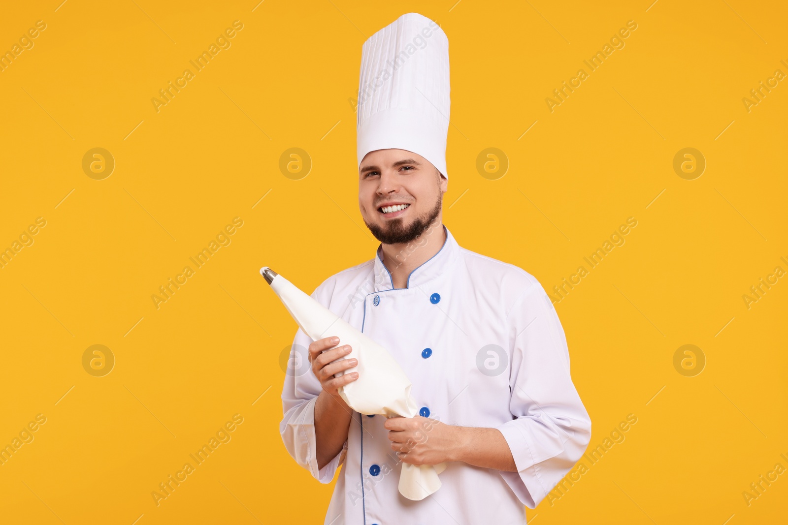 Photo of Happy professional confectioner in uniform holding piping bag on yellow background