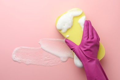 Photo of Cleaner in rubber glove holding sponge with foam on pink background, top view.