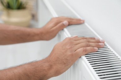 Photo of Man warming hands on heating radiator near white wall, closeup