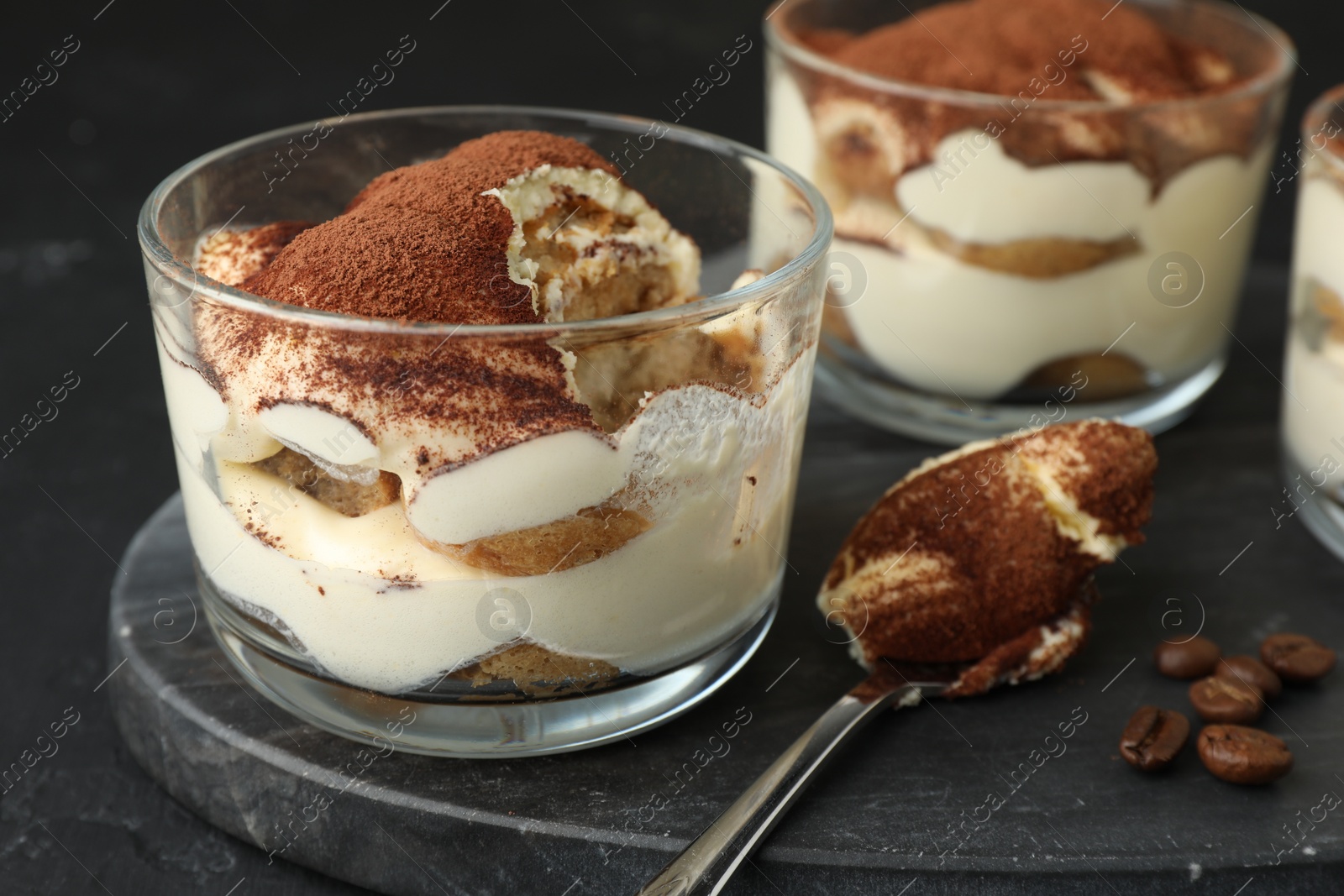 Photo of Delicious tiramisu in glasses, spoon and coffee beans on black table, closeup