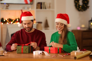 Photo of Happy couple in Santa hats making Christmas gifts at table in room