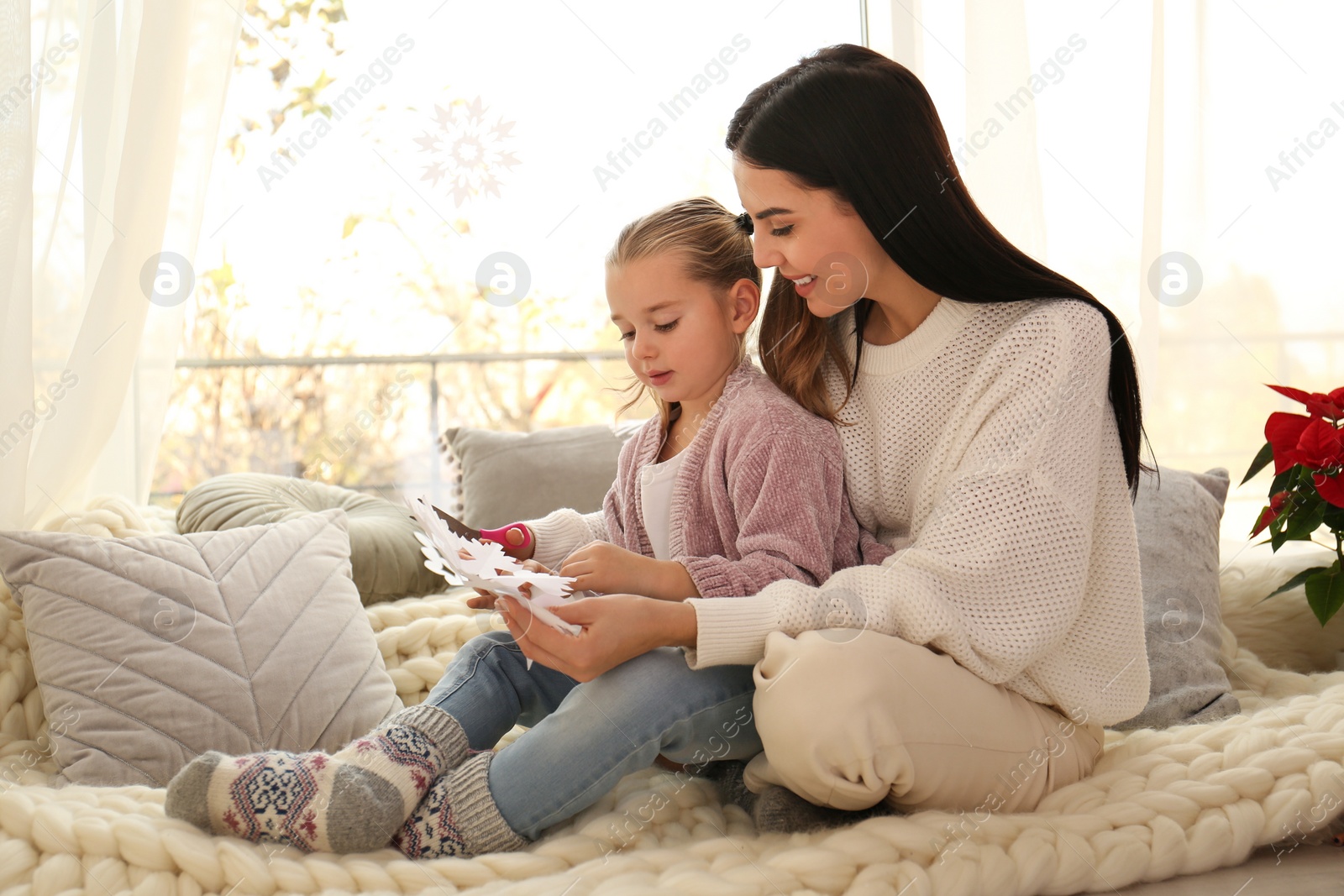 Photo of Mother and daughter making paper snowflake near window at home