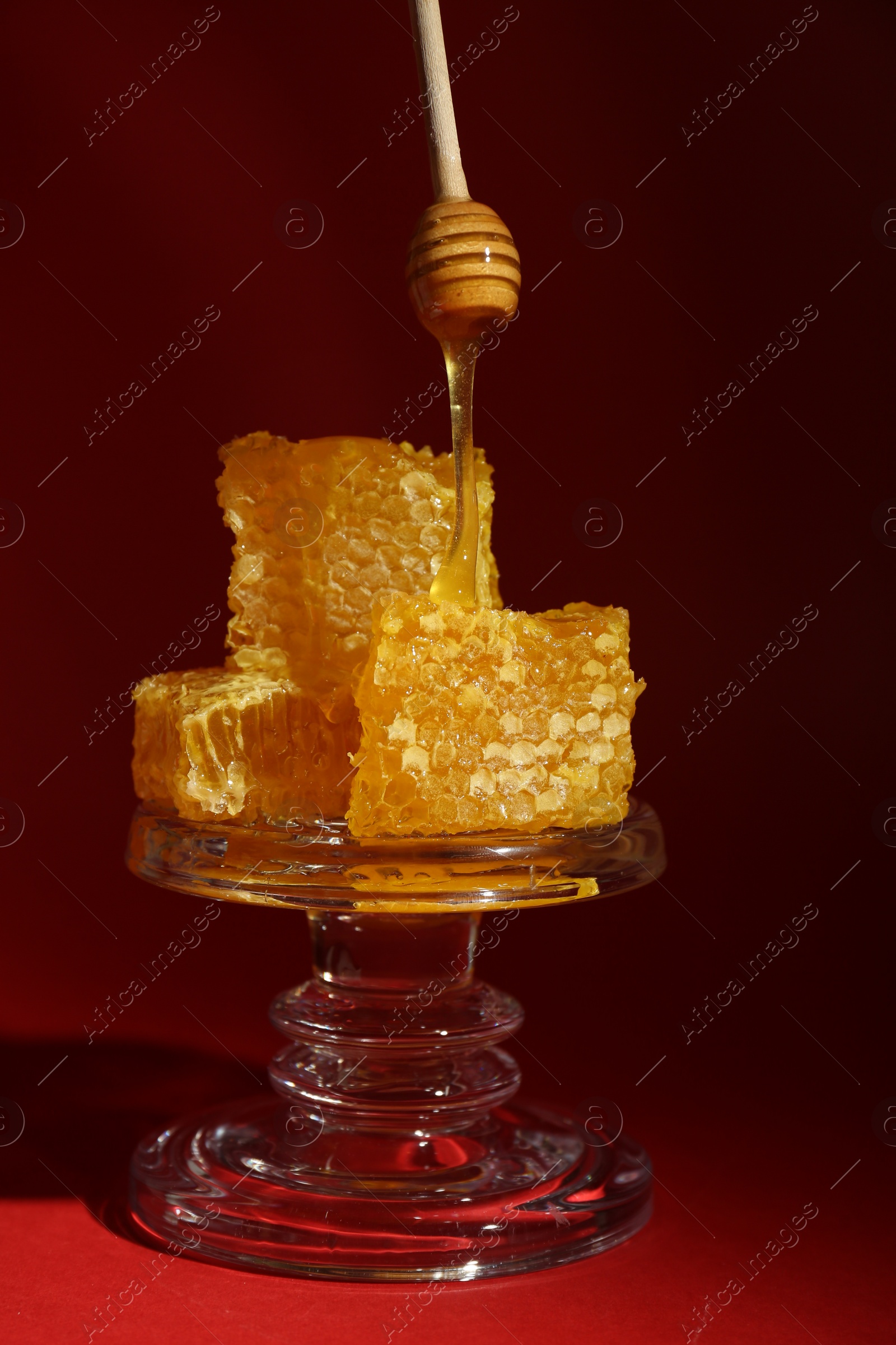 Photo of Dripping tasty honey from dipper onto honeycombs on glass stand against burgundy background