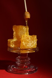 Photo of Dripping tasty honey from dipper onto honeycombs on glass stand against burgundy background