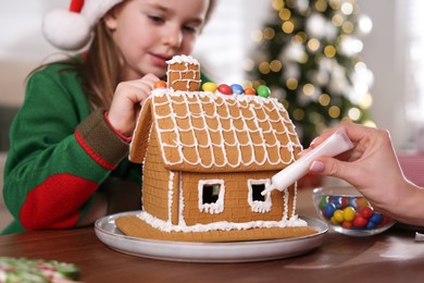 Photo of Mother and daughter decorating gingerbread house at table indoors, closeup