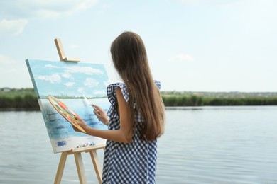 Photo of Little girl painting scenery on easel near lake, back view