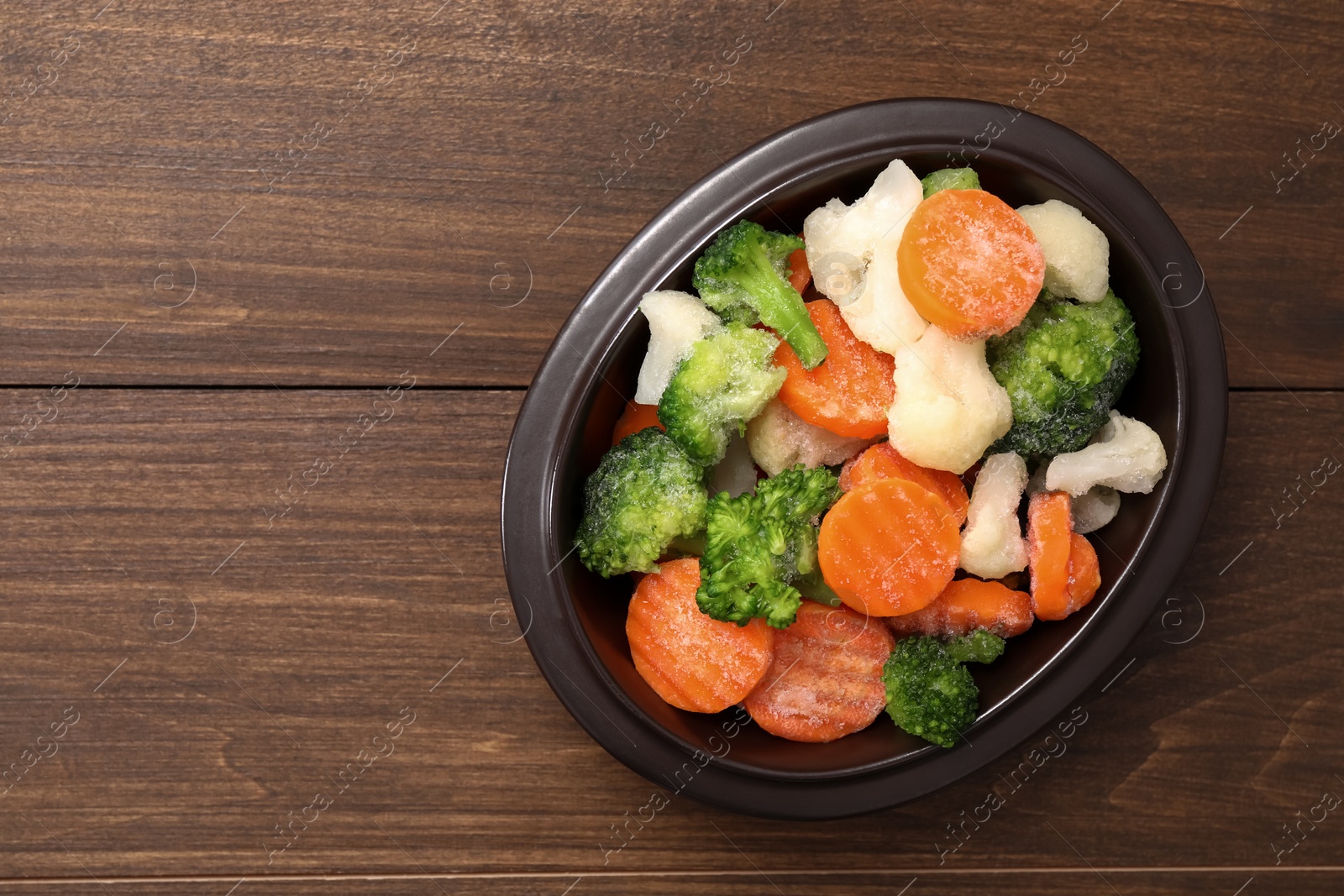 Photo of Mix of different frozen vegetables in bowl on wooden table, top view. Space for text