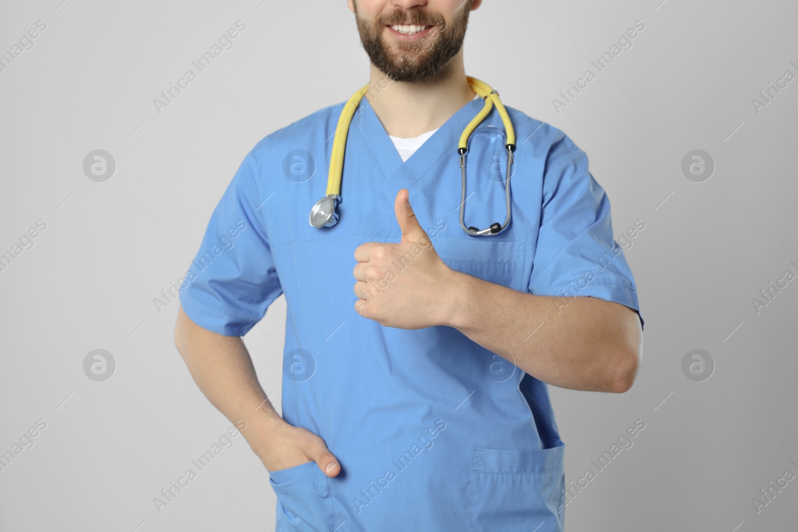 Photo of Doctor or medical assistant (male nurse) with stethoscope showing thumb up on light grey background, closeup