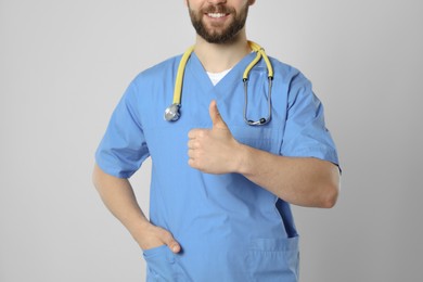 Doctor or medical assistant (male nurse) with stethoscope showing thumb up on light grey background, closeup