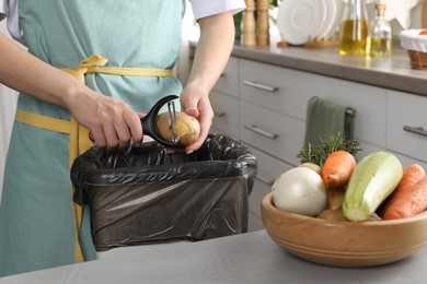 Photo of Woman peeling fresh potato above garbage bin indoors, closeup