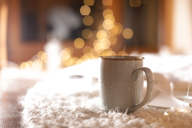 Cup of hot beverage on fuzzy rug against blurred background, space for text. Winter evening