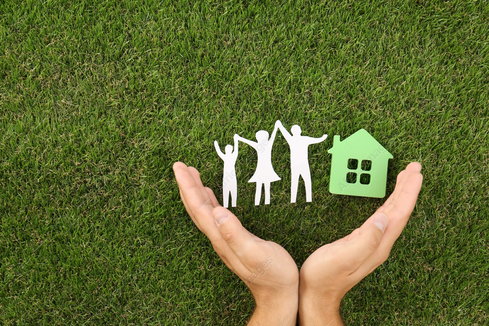 Photo of Man holding hands near figures of house and family on green grass, top view. Space for text