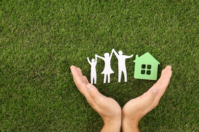Photo of Man holding hands near figures of house and family on green grass, top view. Space for text