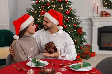 Beautiful young woman in Santa hat presenting gift to her boyfriend at table served for Christmas