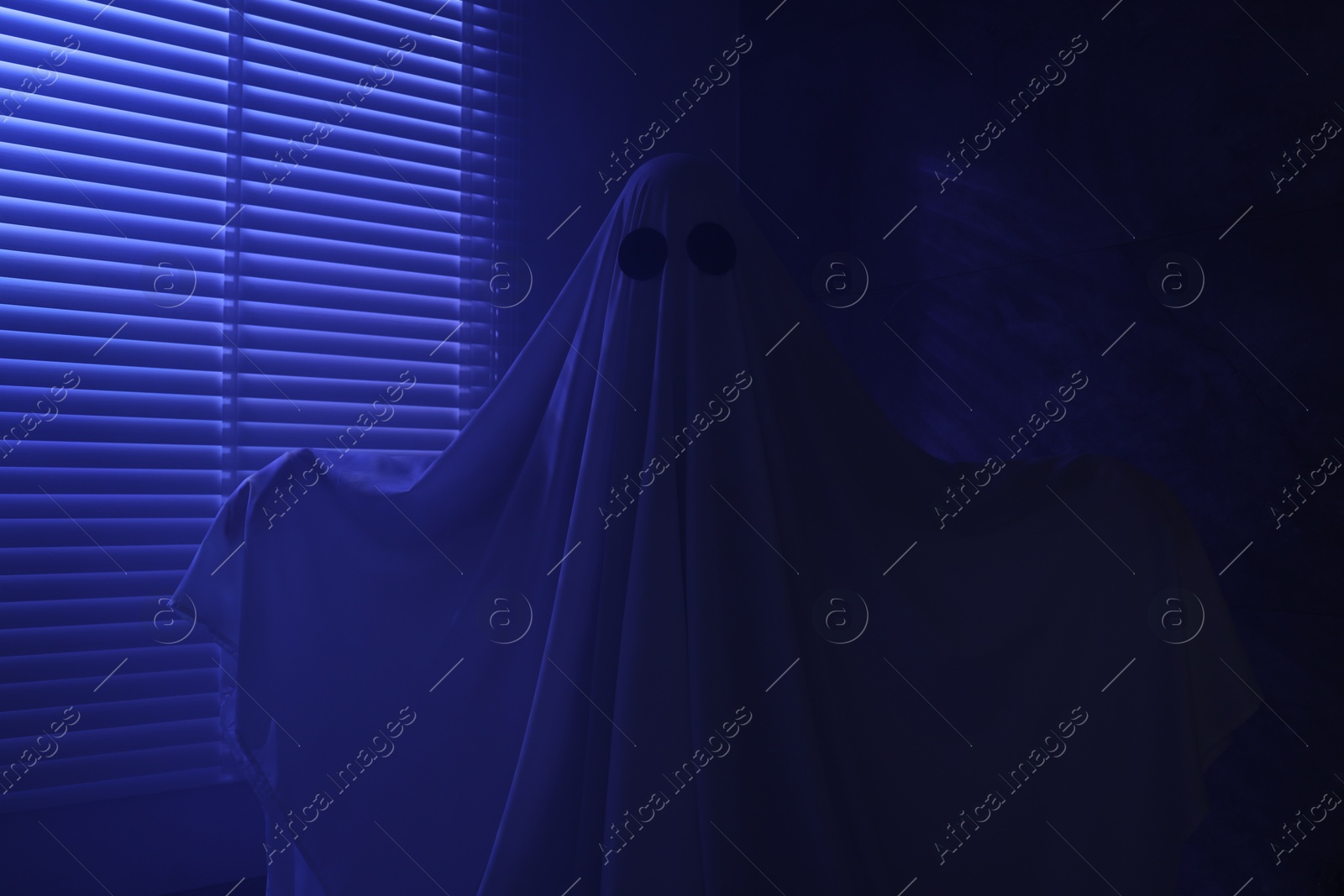 Photo of Creepy ghost. Woman covered with sheet near window in blue light