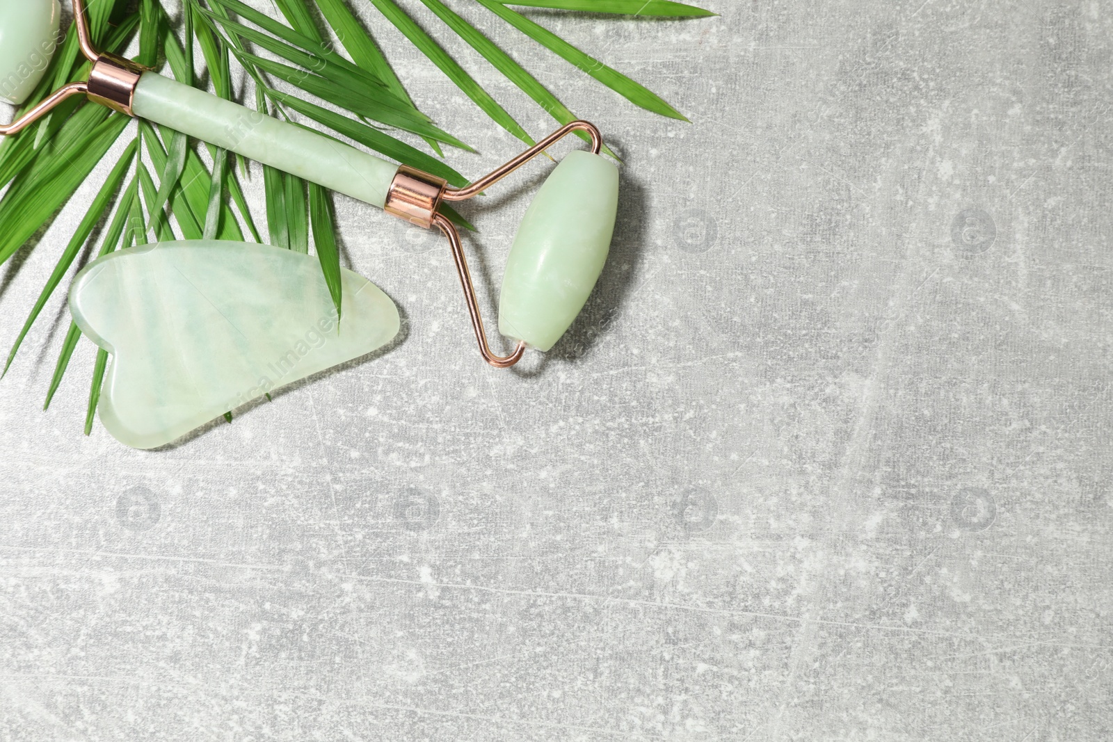 Photo of Quartz gua sha tool, face roller and green leaves on grey background, flat lay. Space for text