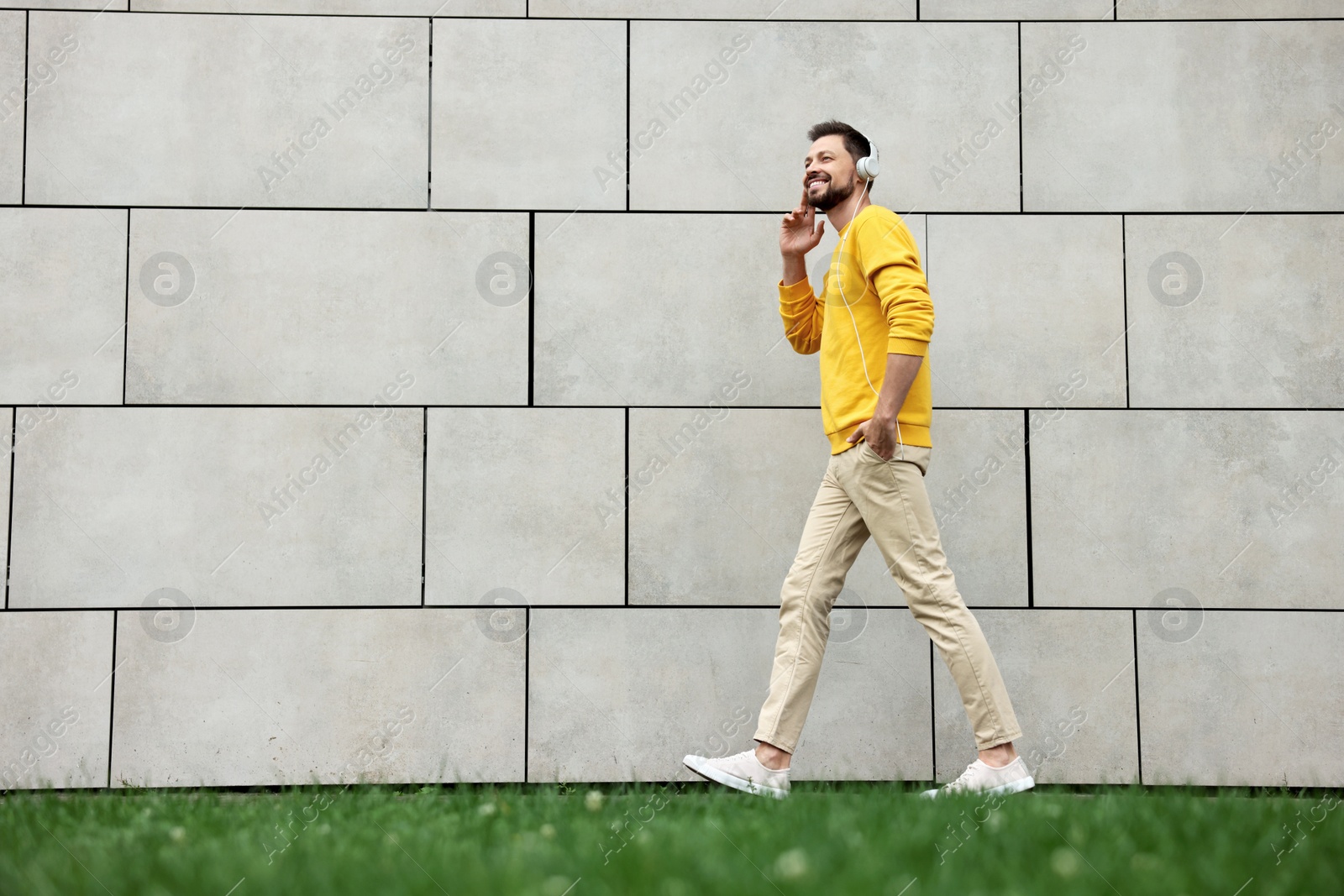 Photo of Handsome man with headphones walking near grey stone wall outdoors, space for text