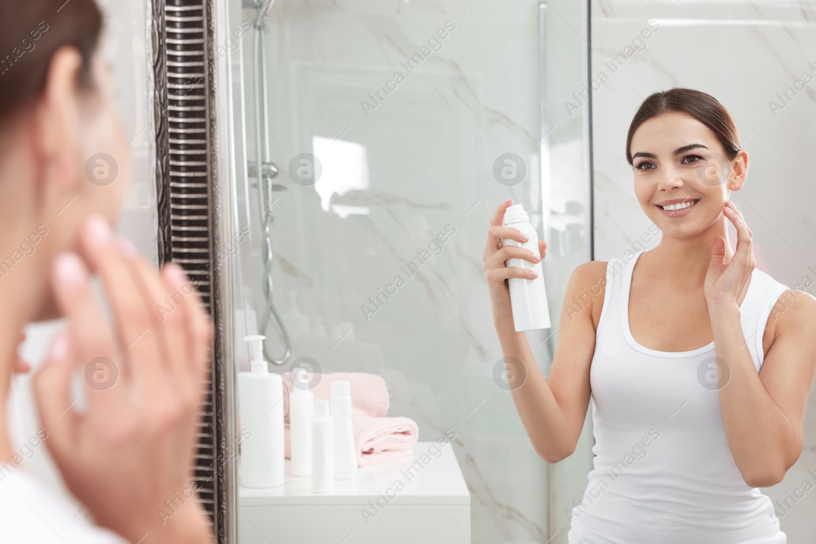 Photo of Young woman applying thermal water on face near mirror in bathroom. Cosmetic product