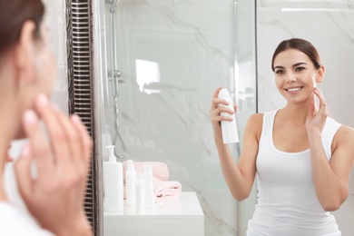 Young woman applying thermal water on face near mirror in bathroom. Cosmetic product