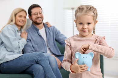 Photo of Family budget. Little girl putting coin into piggy bank while her parents watching indoors, selective focus