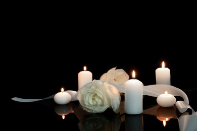 White roses and burning candles on black mirror surface in darkness, space for text. Funeral symbols