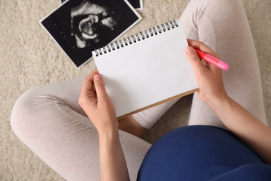 Photo of Pregnant woman with notebook and sonogram choosing name for baby on carpet, top view