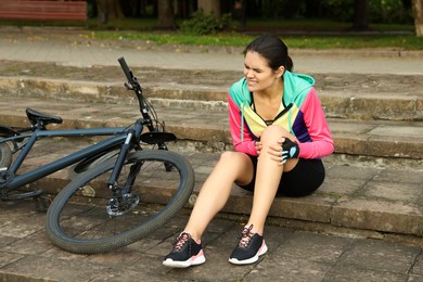 Photo of Young woman with injured knee on steps near bicycle outdoors