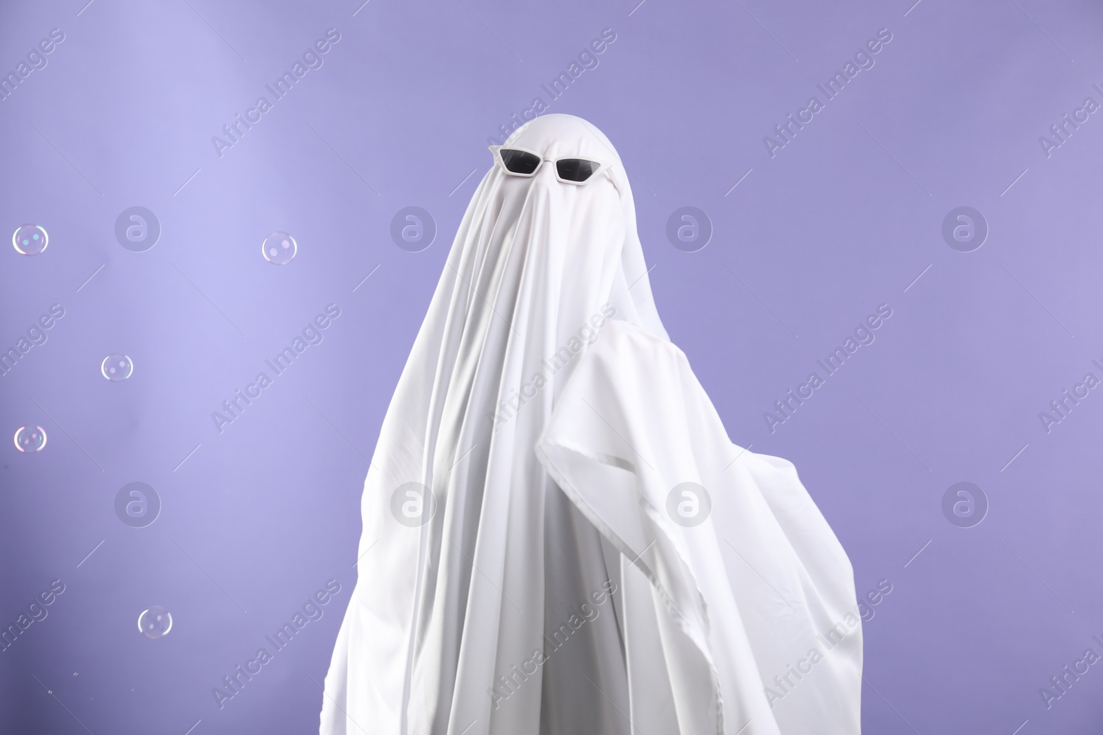 Photo of Funny ghost. Person in white sheet, sunglasses and soap bubbles on violet background