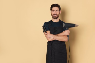 Photo of Smiling hairdresser in apron holding dryer and brush on light brown background, space for text