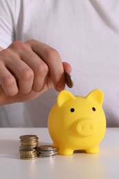 Man putting coin into yellow piggy bank at white table, closeup