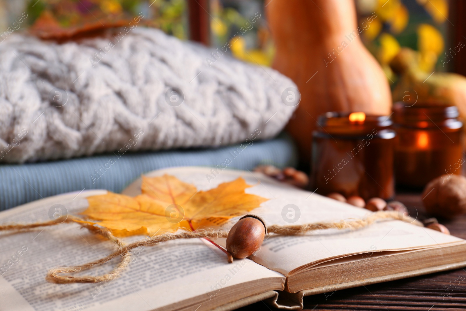 Photo of Book with autumn leaf as bookmark, acorns, scented candle and warm sweaters on wooden table, closeup