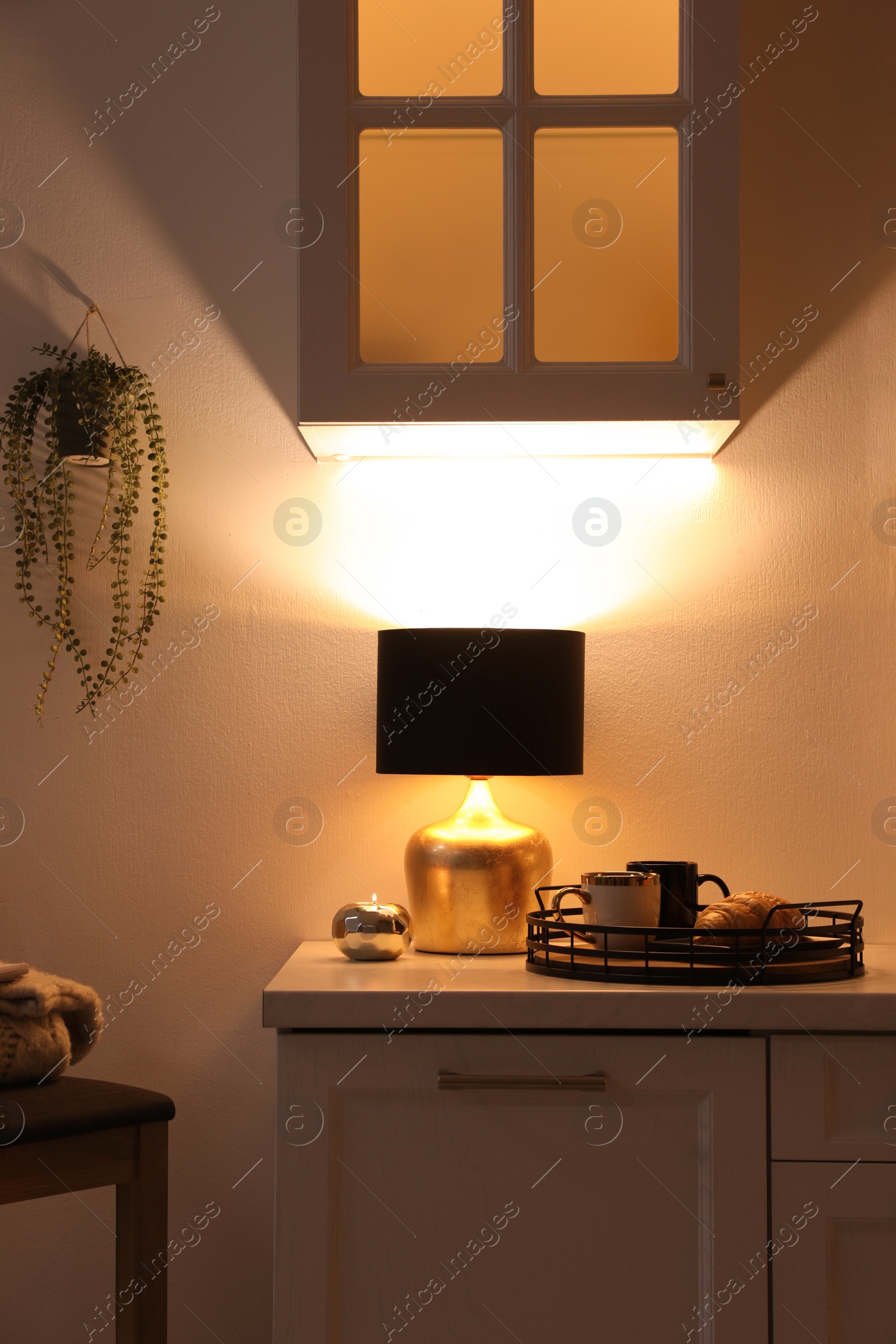 Photo of Stylish lamp, cups and croissant on white cabinet in room. Interior element