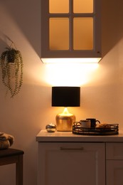 Stylish lamp, cups and croissant on white cabinet in room. Interior element