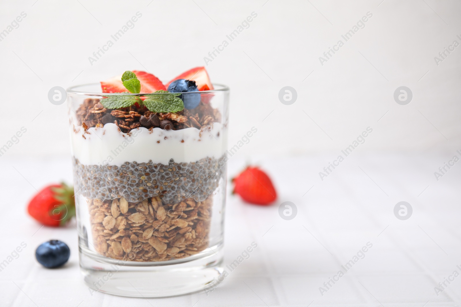 Photo of Tasty granola with berries, yogurt and chia seeds in glass on white tiled table, space for text