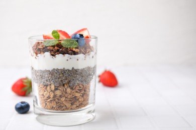 Photo of Tasty granola with berries, yogurt and chia seeds in glass on white tiled table, space for text