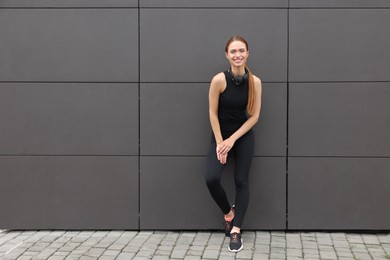 Photo of Beautiful woman in stylish gym clothes with headphones near dark grey wall on street, space for text