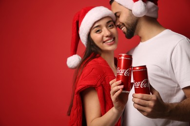 Photo of MYKOLAIV, UKRAINE - JANUARY 27, 2021: Young couple in Christmas hats holding cans of Coca-Cola against red background, focus on hands. Space for text