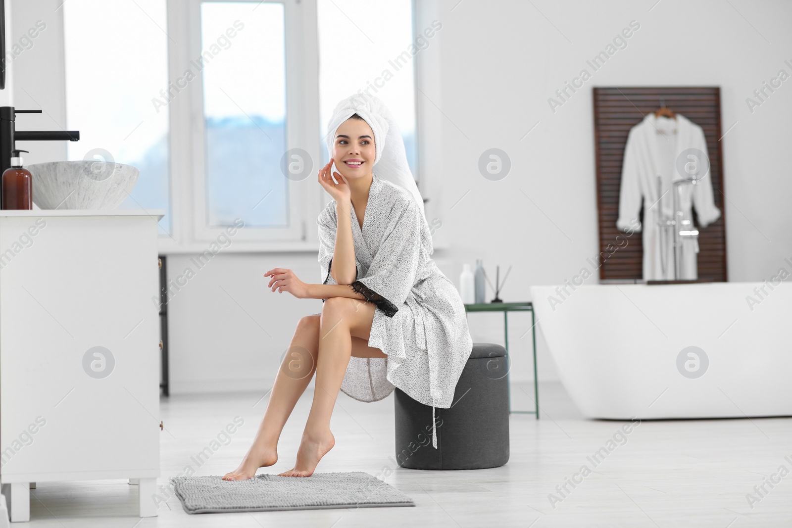 Photo of Beautiful happy woman in stylish bathrobe sitting on ottoman in bathroom